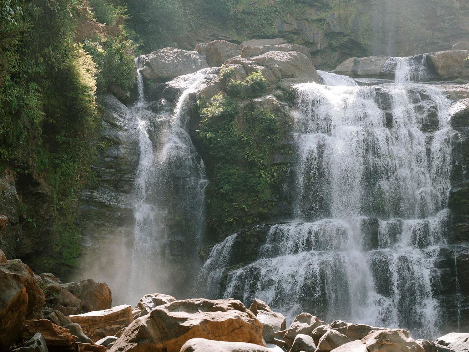 A majestic multi-tiered waterfall cascades down rocky cliffs in a natural forest setting.