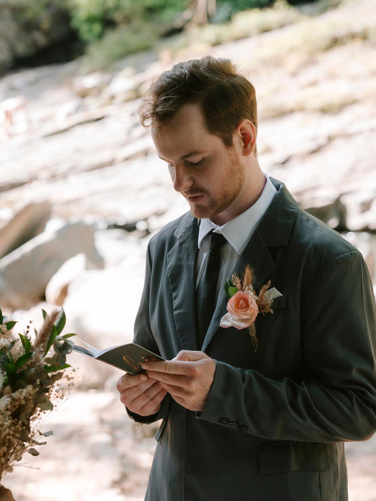 Groom in dark suit reading from small card during outdoor ceremony.