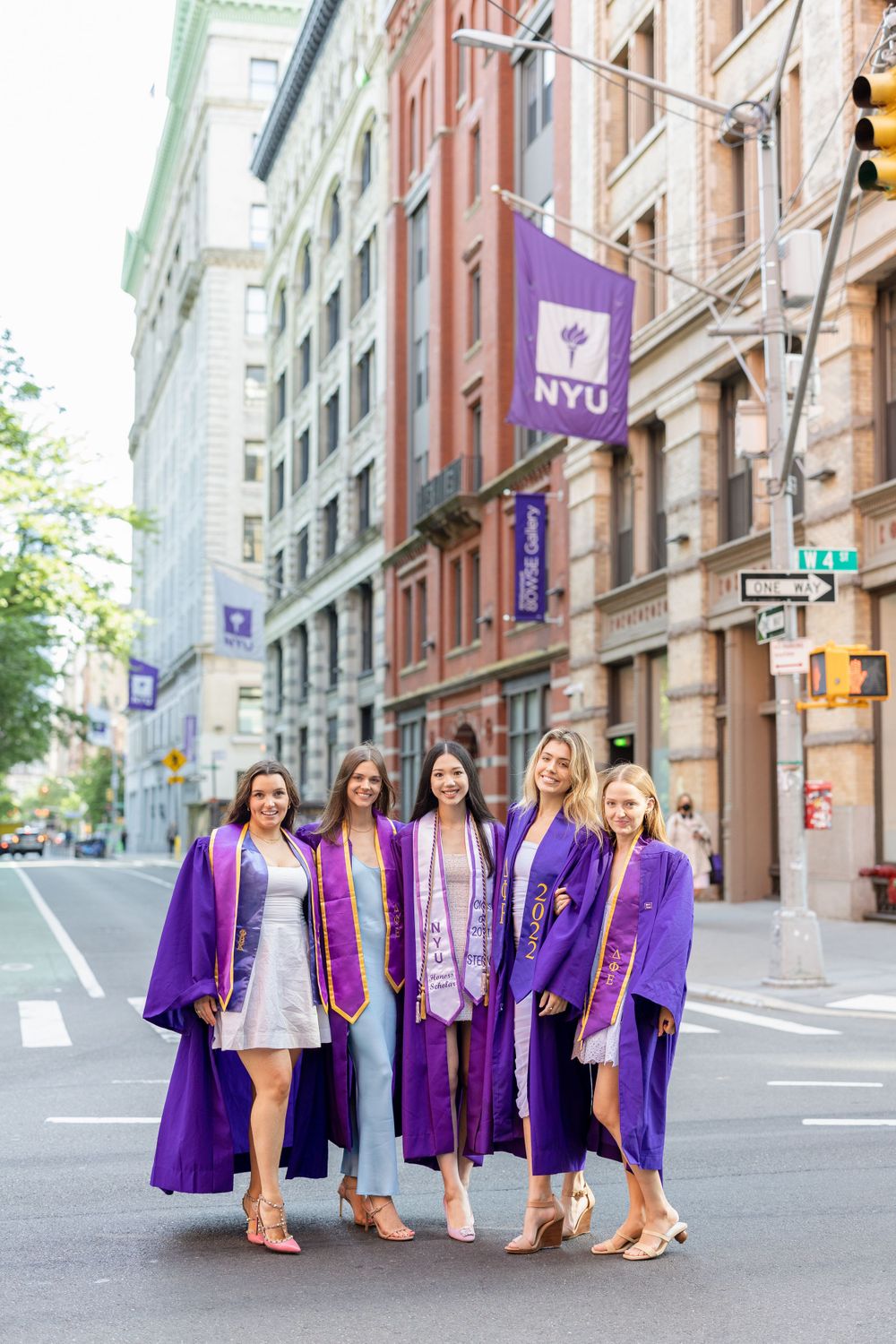 Graduates in purple robes stand together on NYU campus street.
