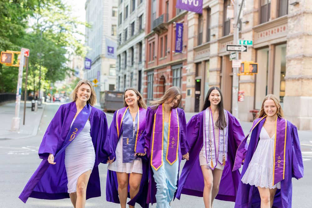 Friends in graduation robes walk together on NYU campus street.