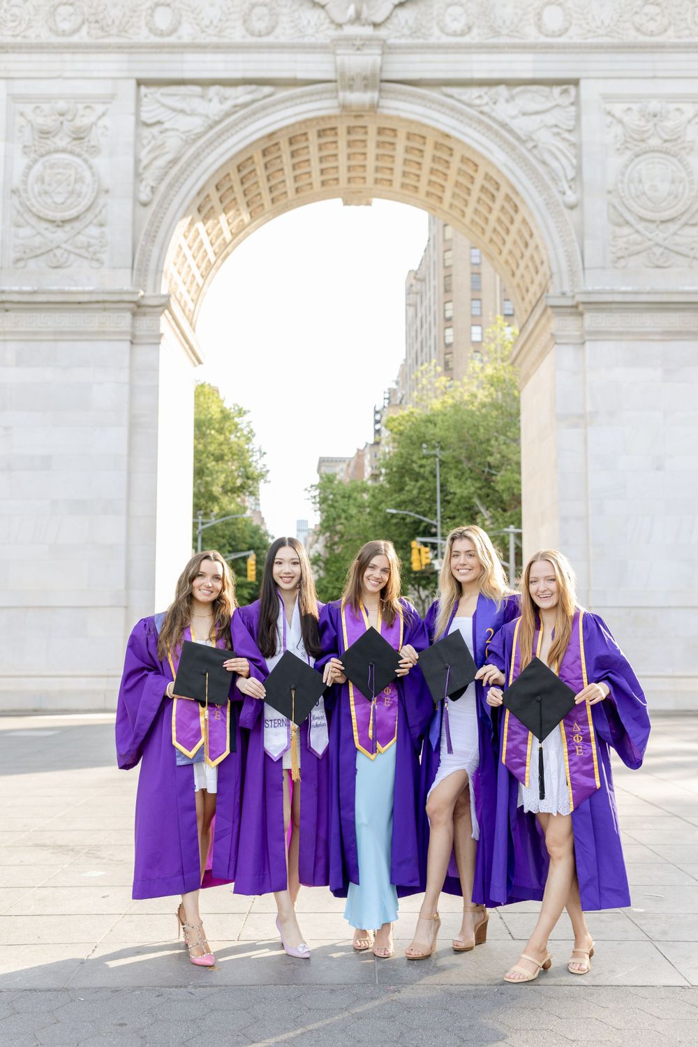 Graduates in purple gowns holding diplomas while standing under Washington Square Arch.