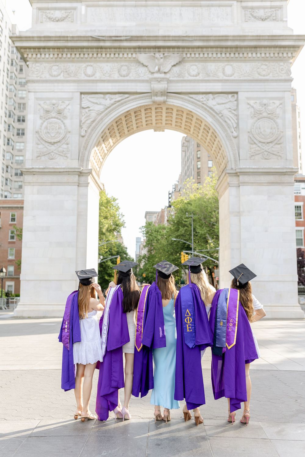 Graduates in purple robes and caps stand facing away under a grand arch.