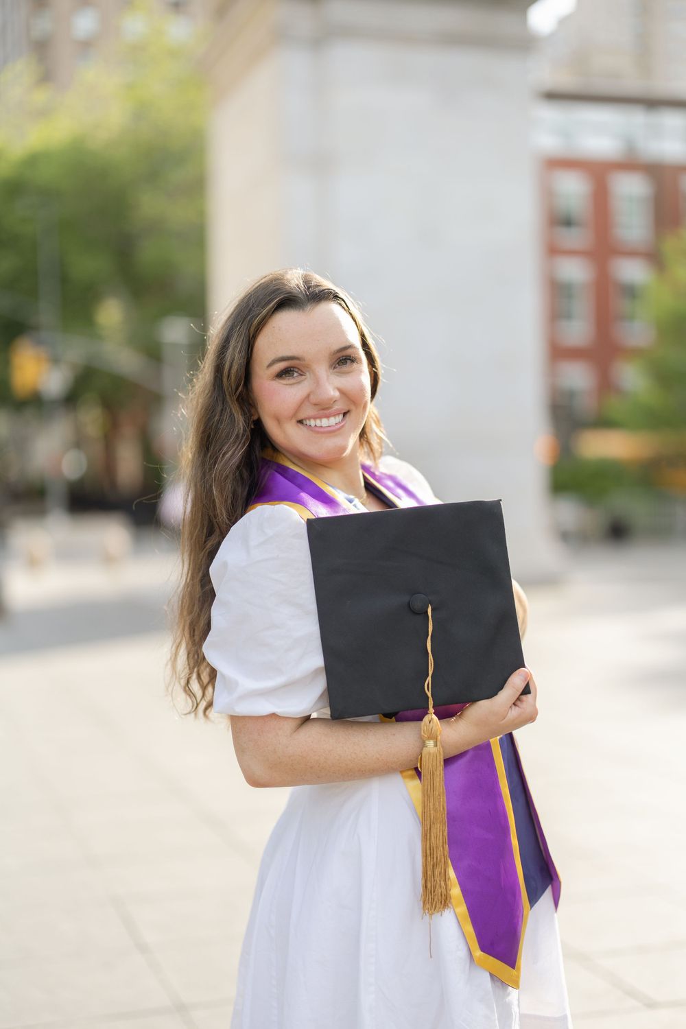 Graduate in white dress holds graduation cap near Washington Square Park.