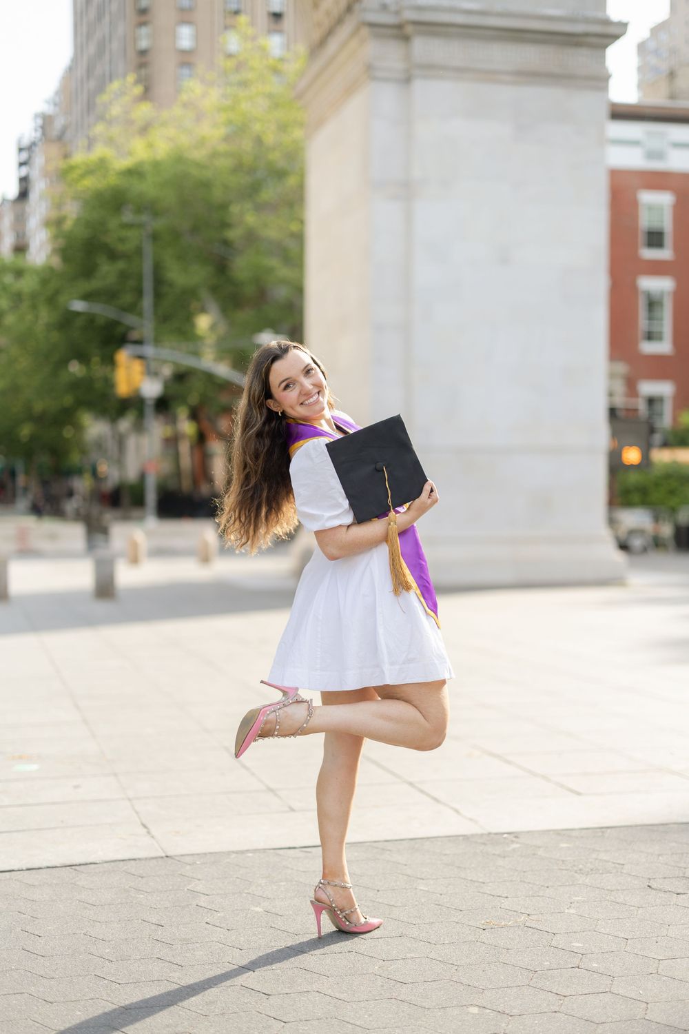 Graduate in white dress twirls with diploma near NYU arch.