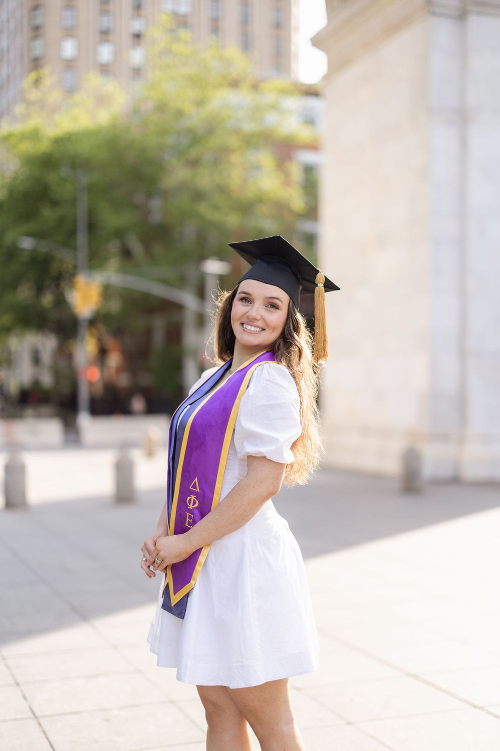 Graduate poses in white dress and purple stole near Washington Square Park.
