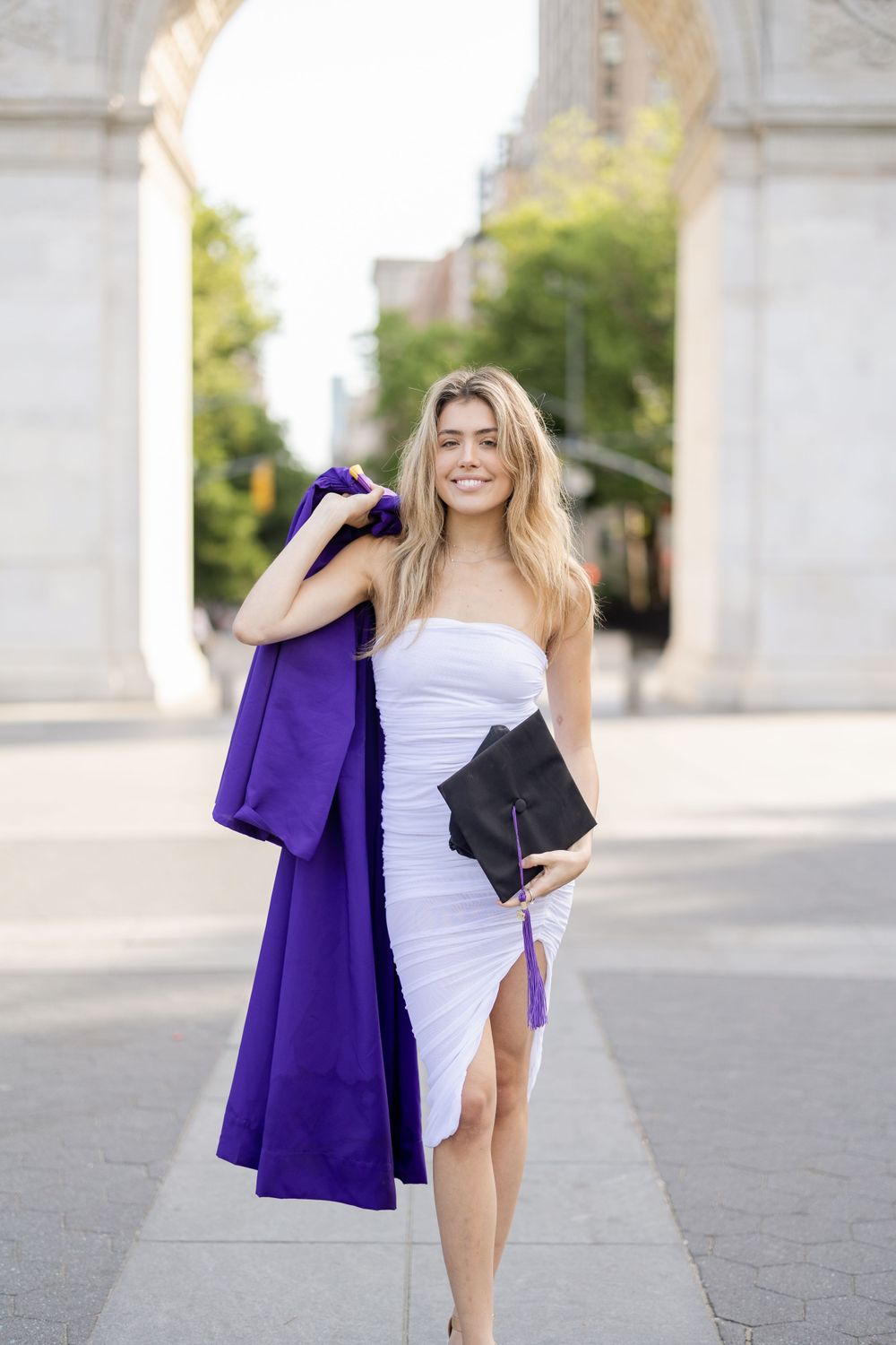 Graduate in white dress carries purple gown and diploma holder near Washington Square Arch.