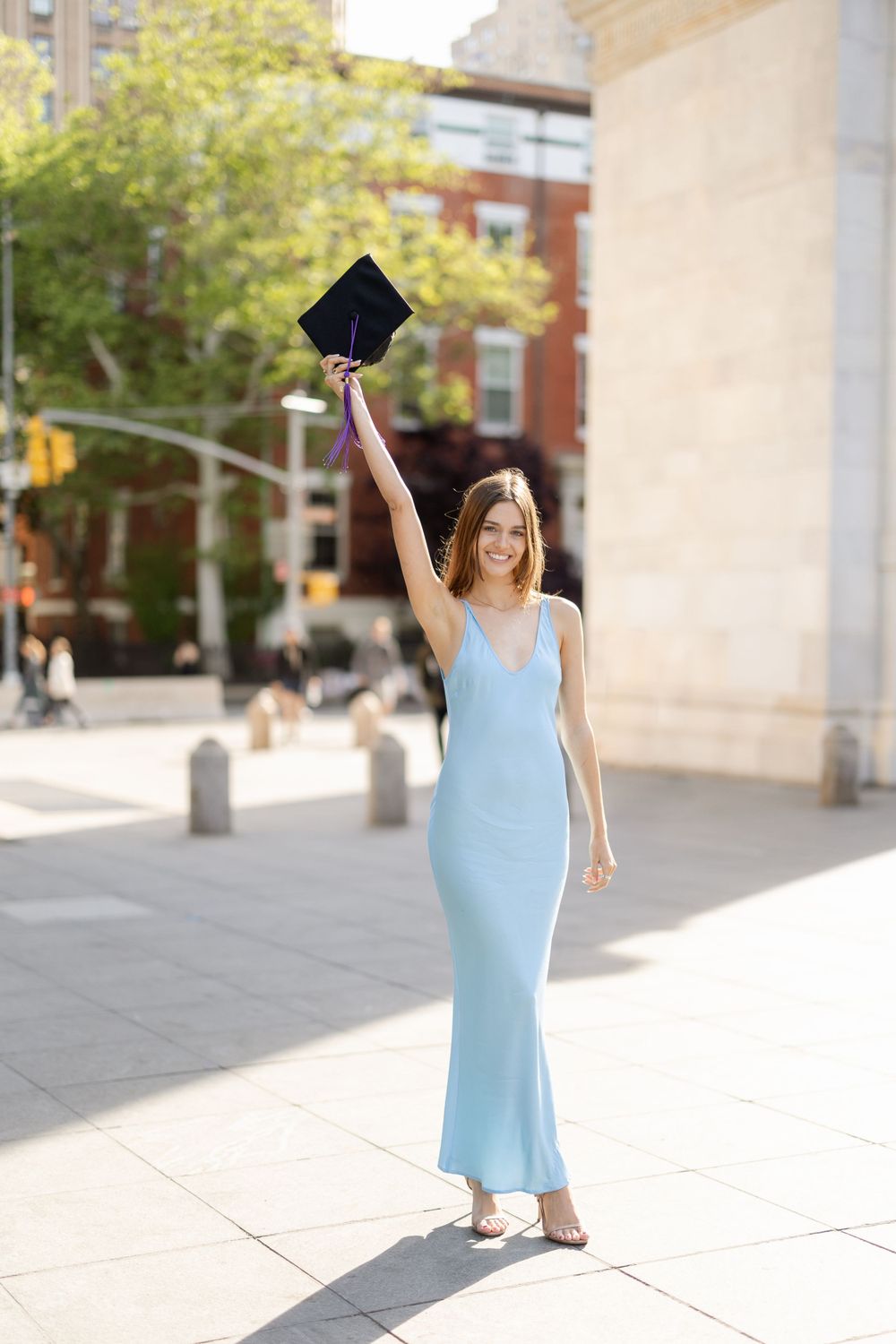 Graduate in blue dress holds diploma holder high near Washington Square Park.