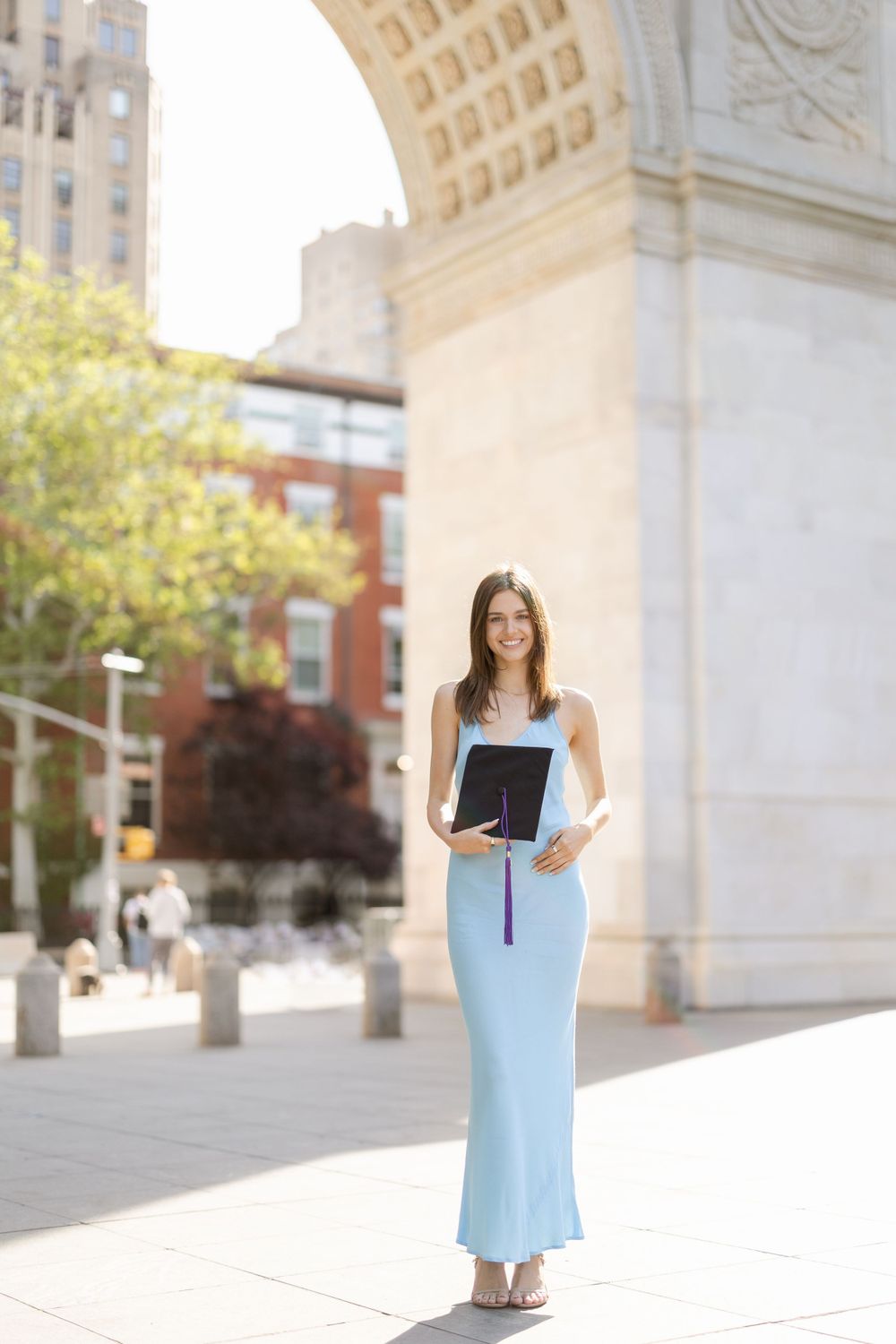 Graduate in light blue dress poses with diploma by Washington Square Arch.
