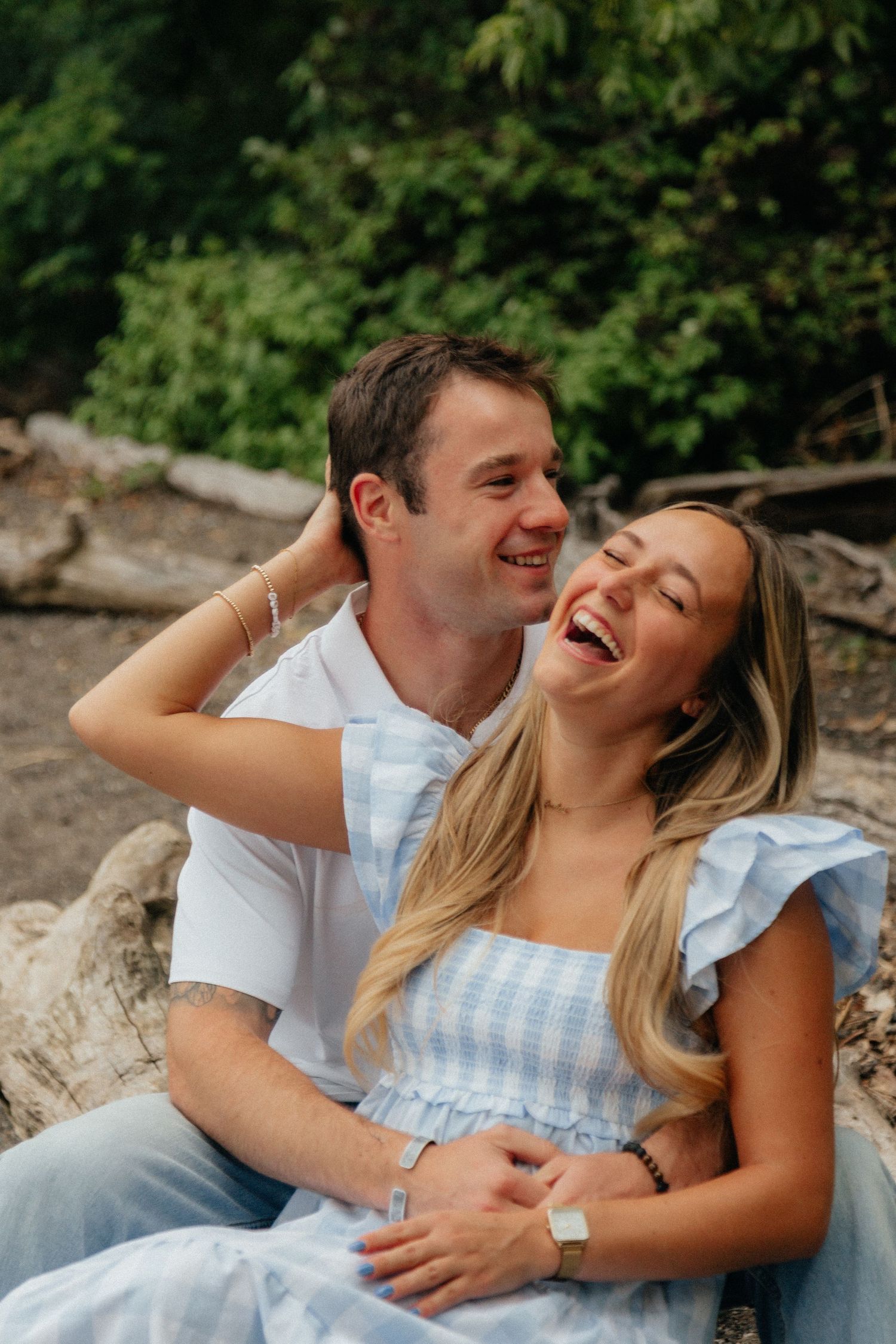 Laughing couple sits together on driftwood outdoors, woman in blue gingham dress, man in white shirt.