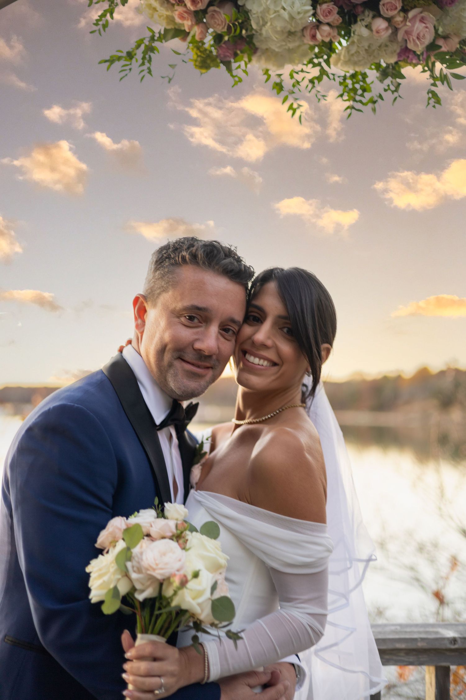 A couple embraces during sunset at a waterfront wedding ceremony under floral decor and warm golden skies.