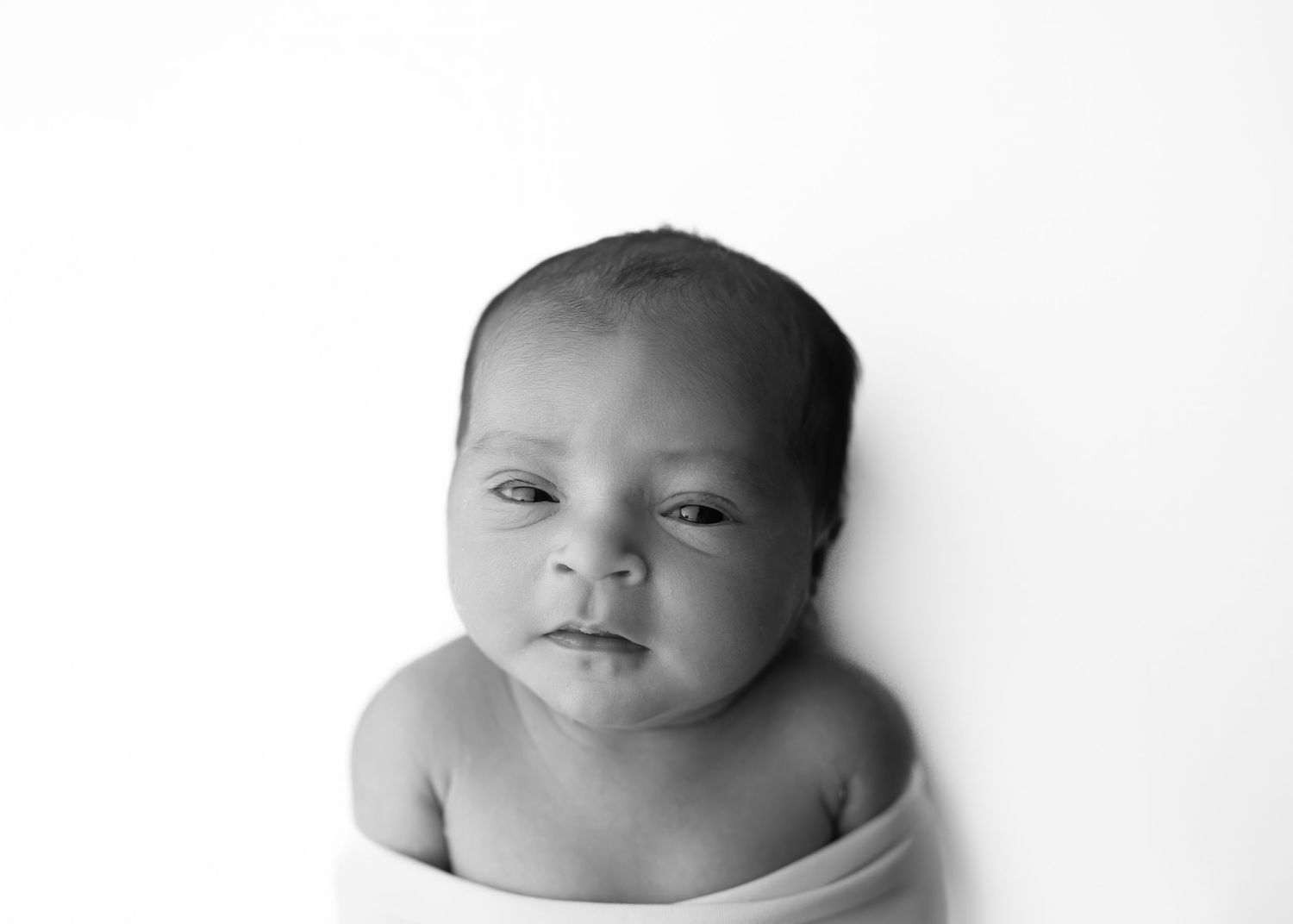 A black and white portrait of a newborn baby swaddled in white fabric looking directly at the camera.