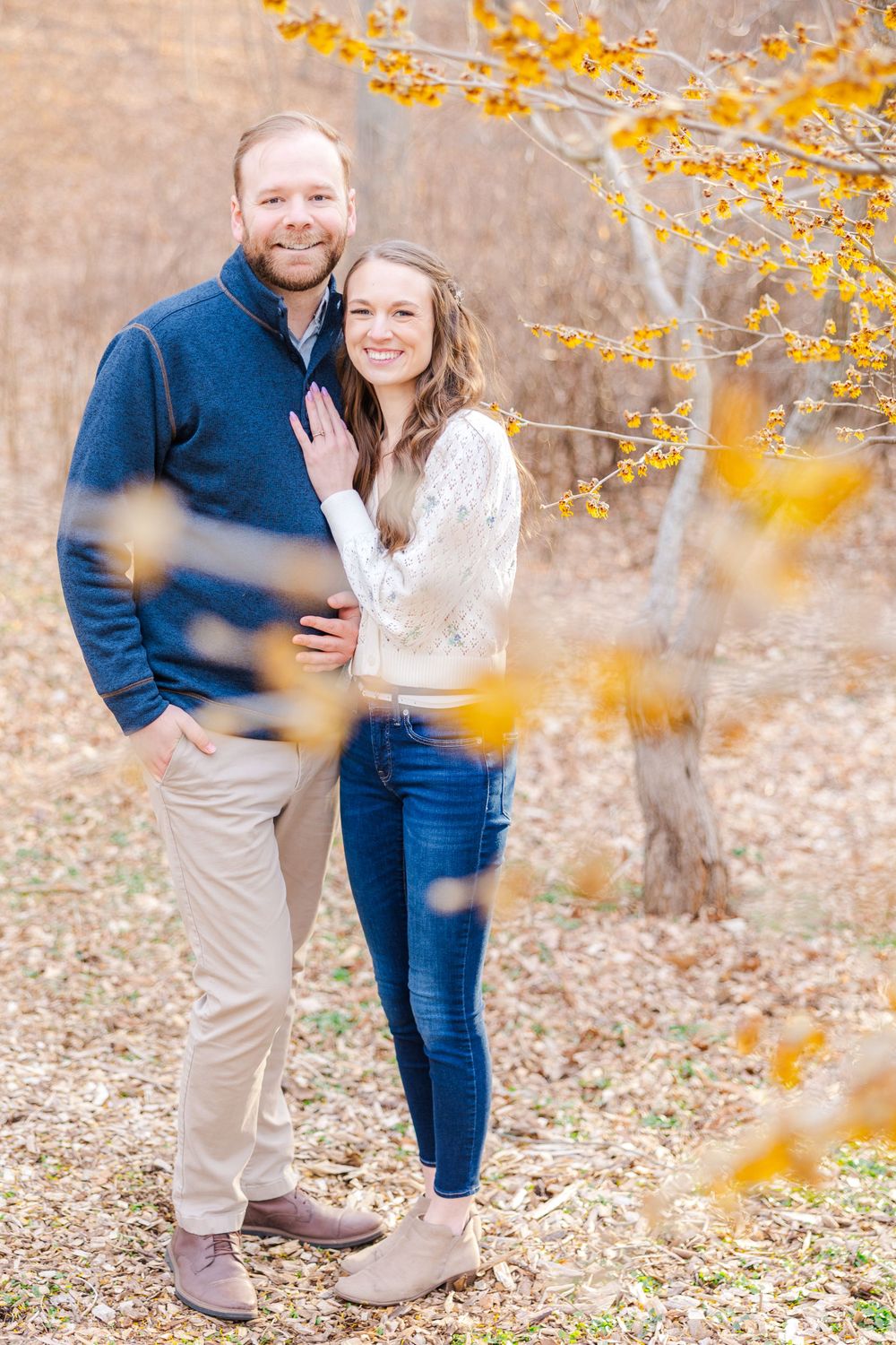 Couple poses together outdoors with yellow autumn branches framing their engagement portrait.