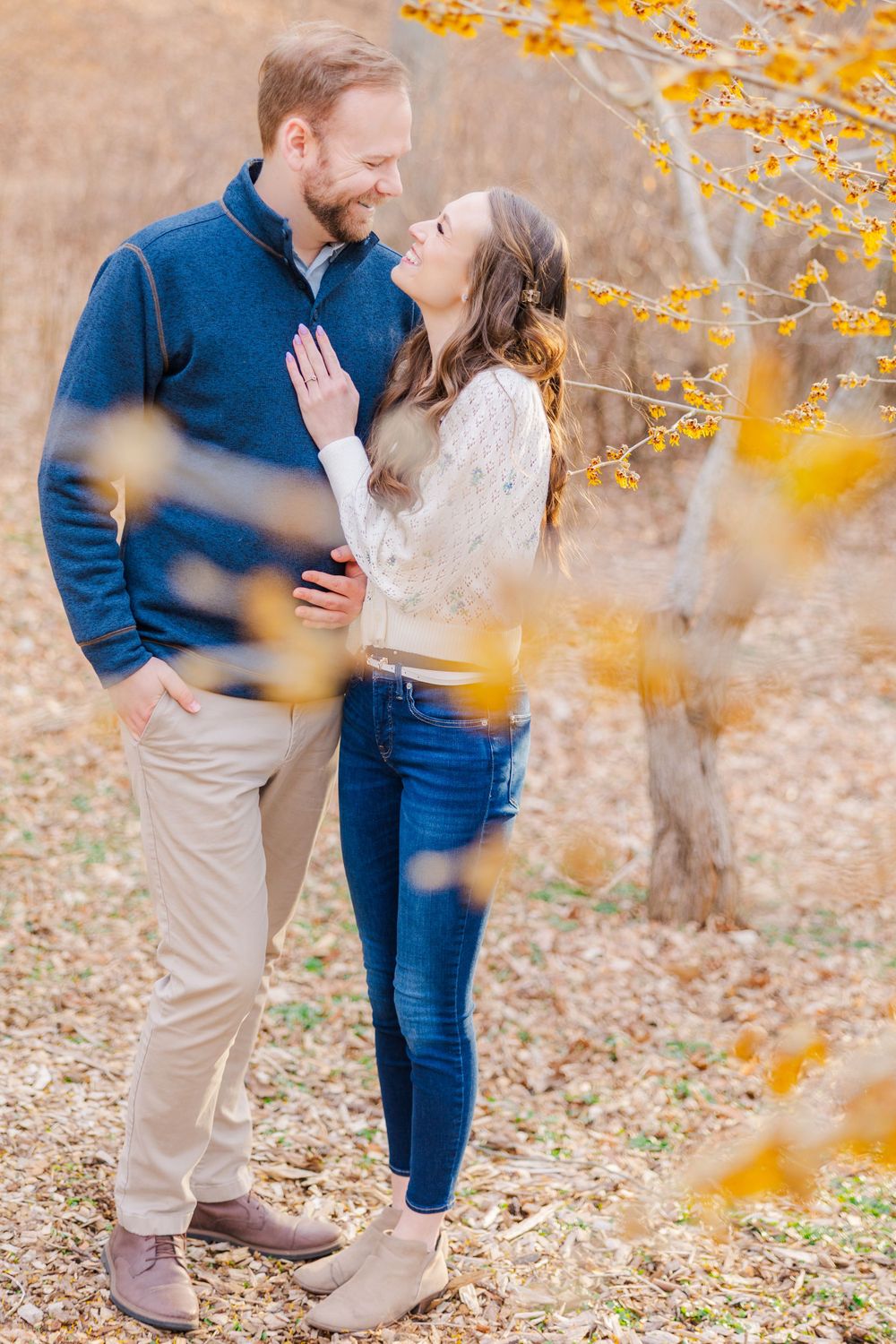 A romantic autumn engagement photo of a couple embracing under yellow fall foliage in a natural outdoor setting.