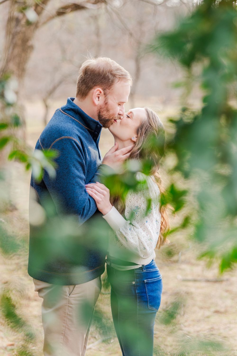 Romantic kiss captured through leaves during an autumn engagement photo session.