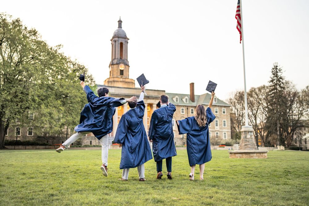 Graduates in blue caps and gowns celebrate by jumping with raised arms on a college campus lawn with flagpole and bell tower.