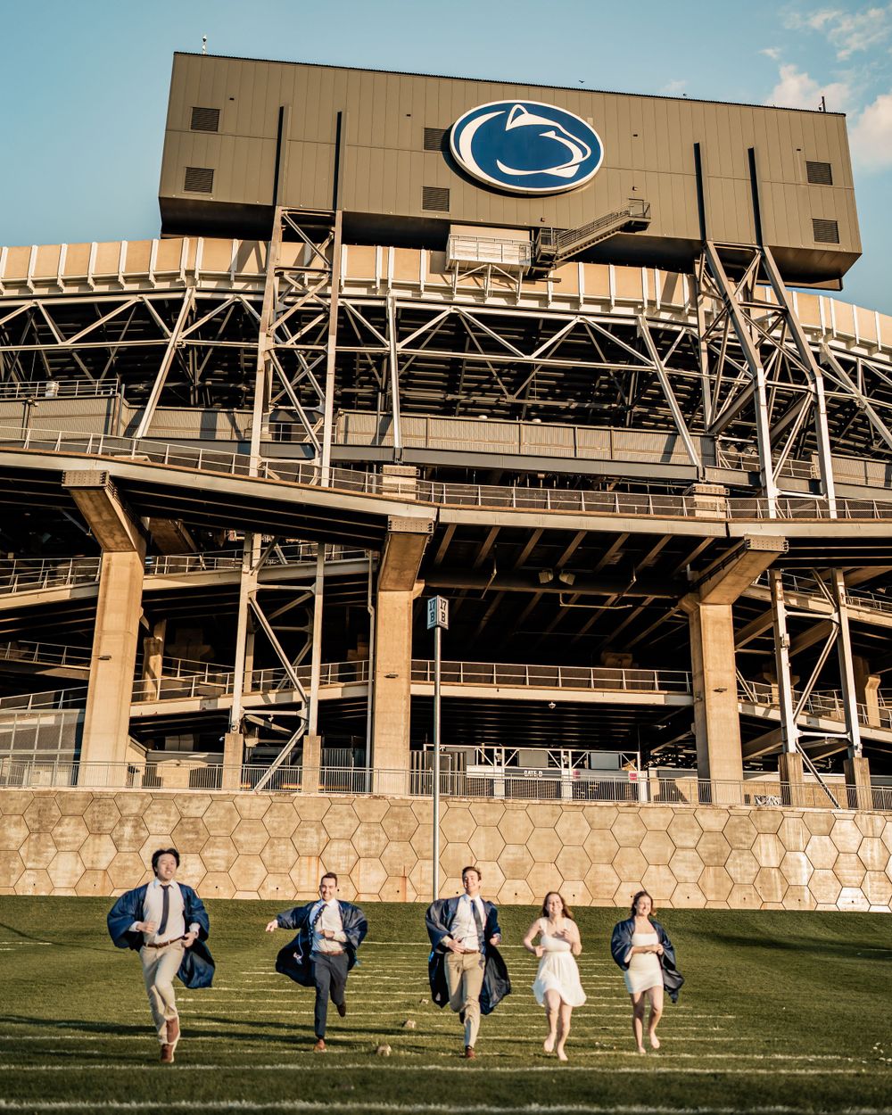 Group runs joyfully across lawn in front of Penn State's Beaver Stadium featuring iconic Nittany Lion logo on a sunny day.