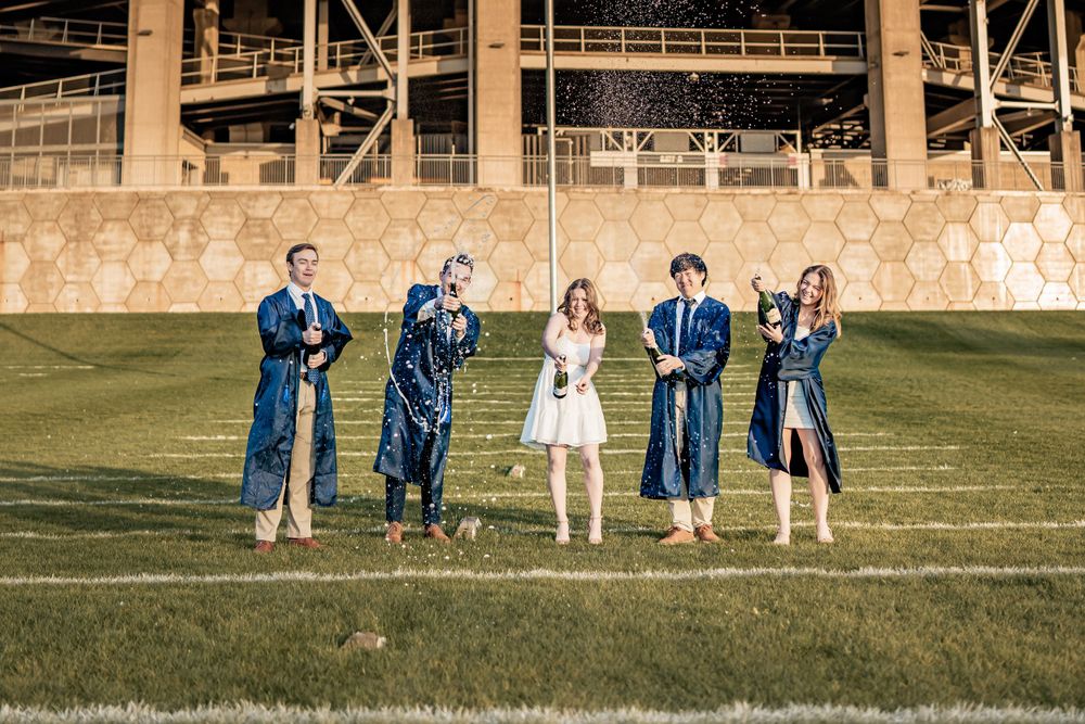 A group in graduation robes and formal attire stand together on a grassy field in front of a sports stadium.