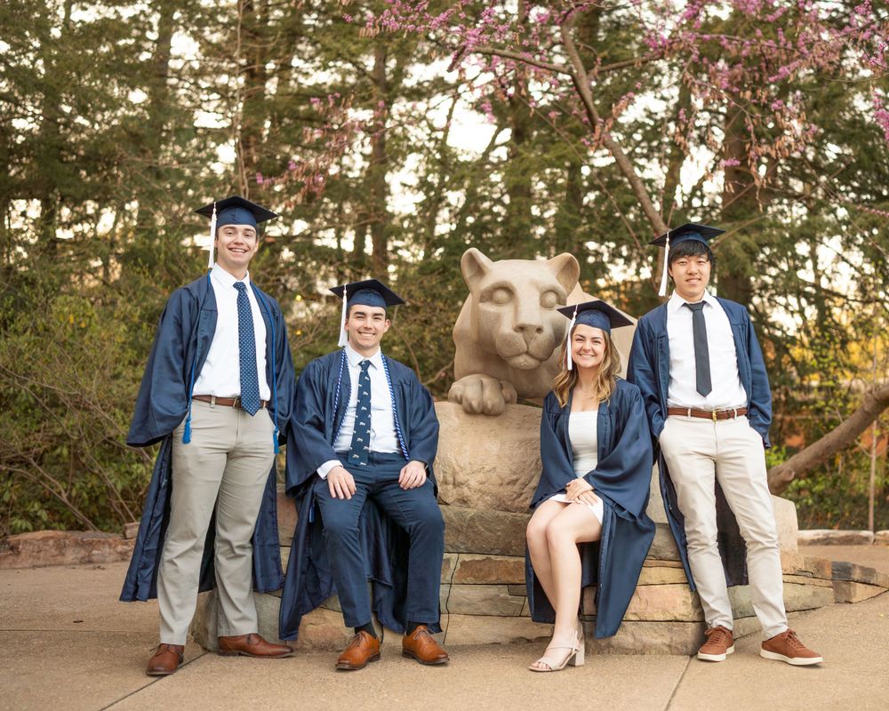 Group of graduates in blue and white attire pose with bronze bear statue on campus grounds during sunset.