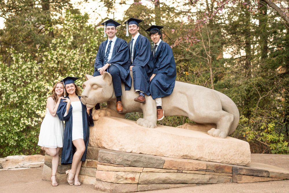 College graduates in blue caps and gowns pose on the Penn State Nittany Lion statue during graduation season.