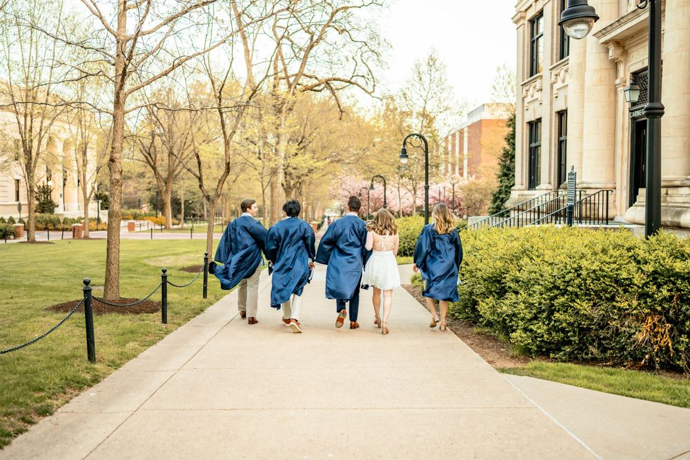 Group in matching blue graduation gowns walk together down a tree-lined campus path on a spring day.