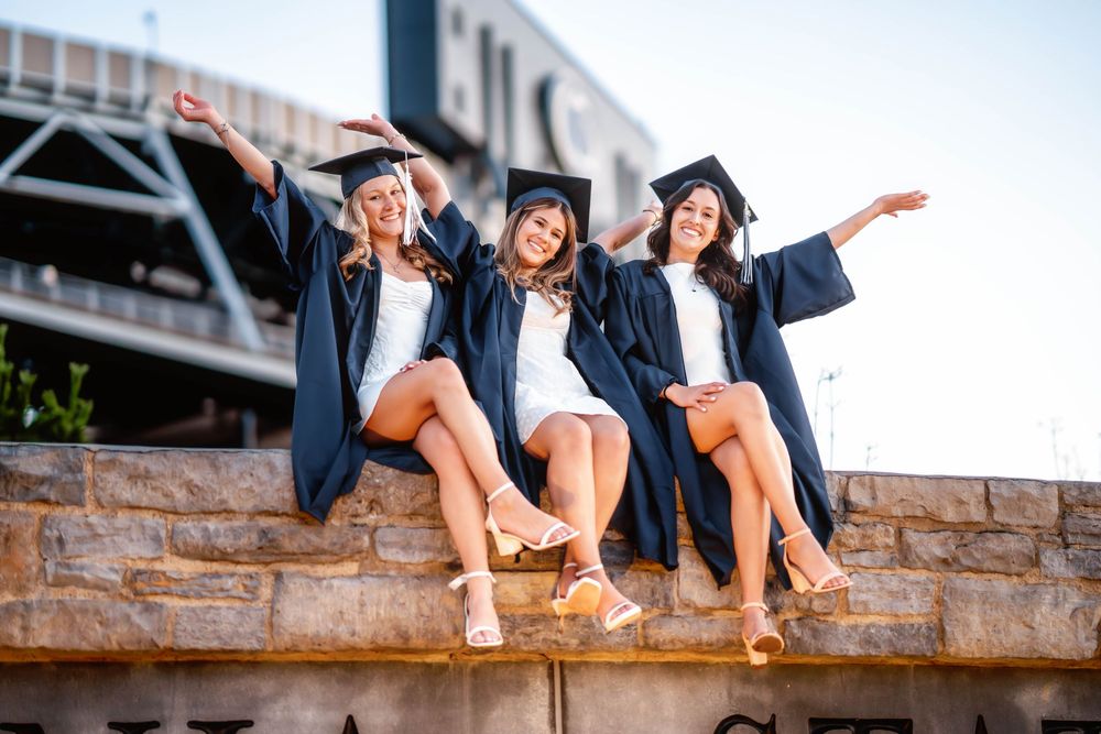 Three happy female graduates in navy caps and gowns sit on a stone wall, arms outstretched, celebrating outdoors.