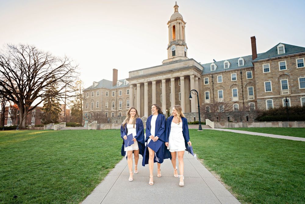 Three female graduates in white dresses and blue gowns walk in front of Penn State's Old Main building.