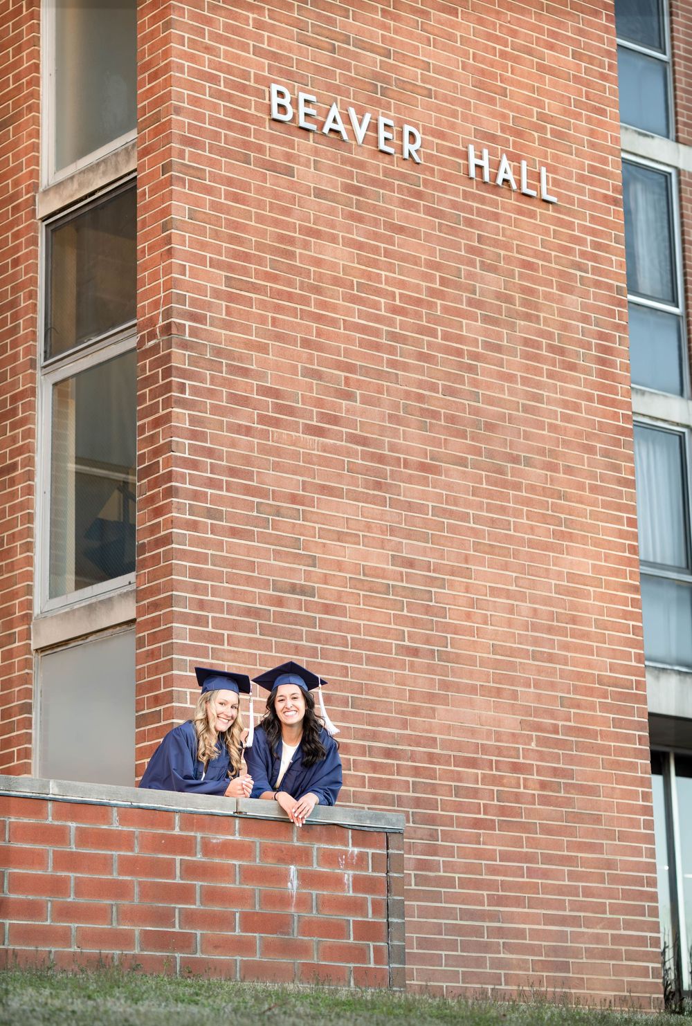 Two graduates in caps and gowns pose at Beaver Hall brick building wall for graduation photos.