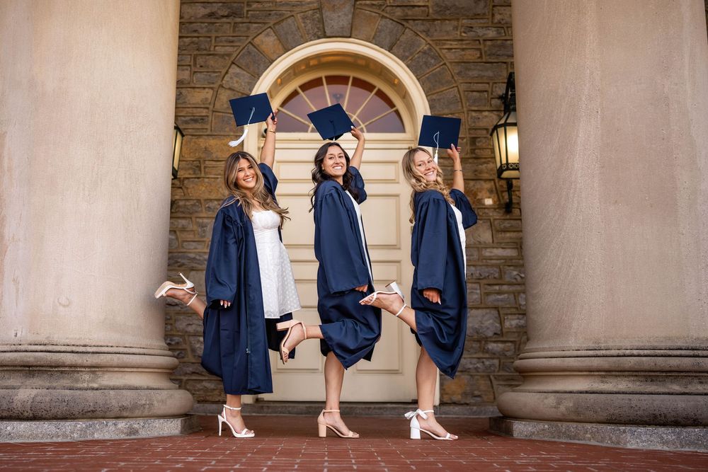 Three women in navy graduation gowns pose joyfully, tossing caps in front of an elegant stone building entrance.