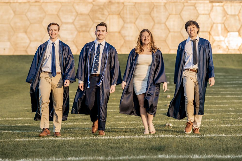 Group of graduates in navy blue gowns walking together on a grassy field during golden hour.