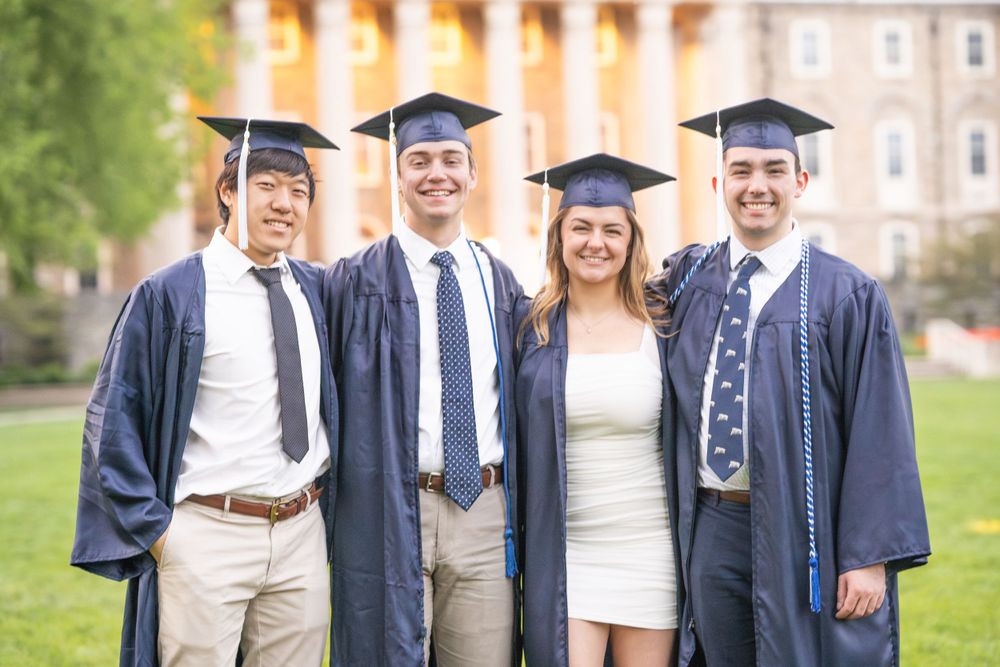 Graduates wearing navy blue commencement robes and mortarboards pose together on campus grounds.