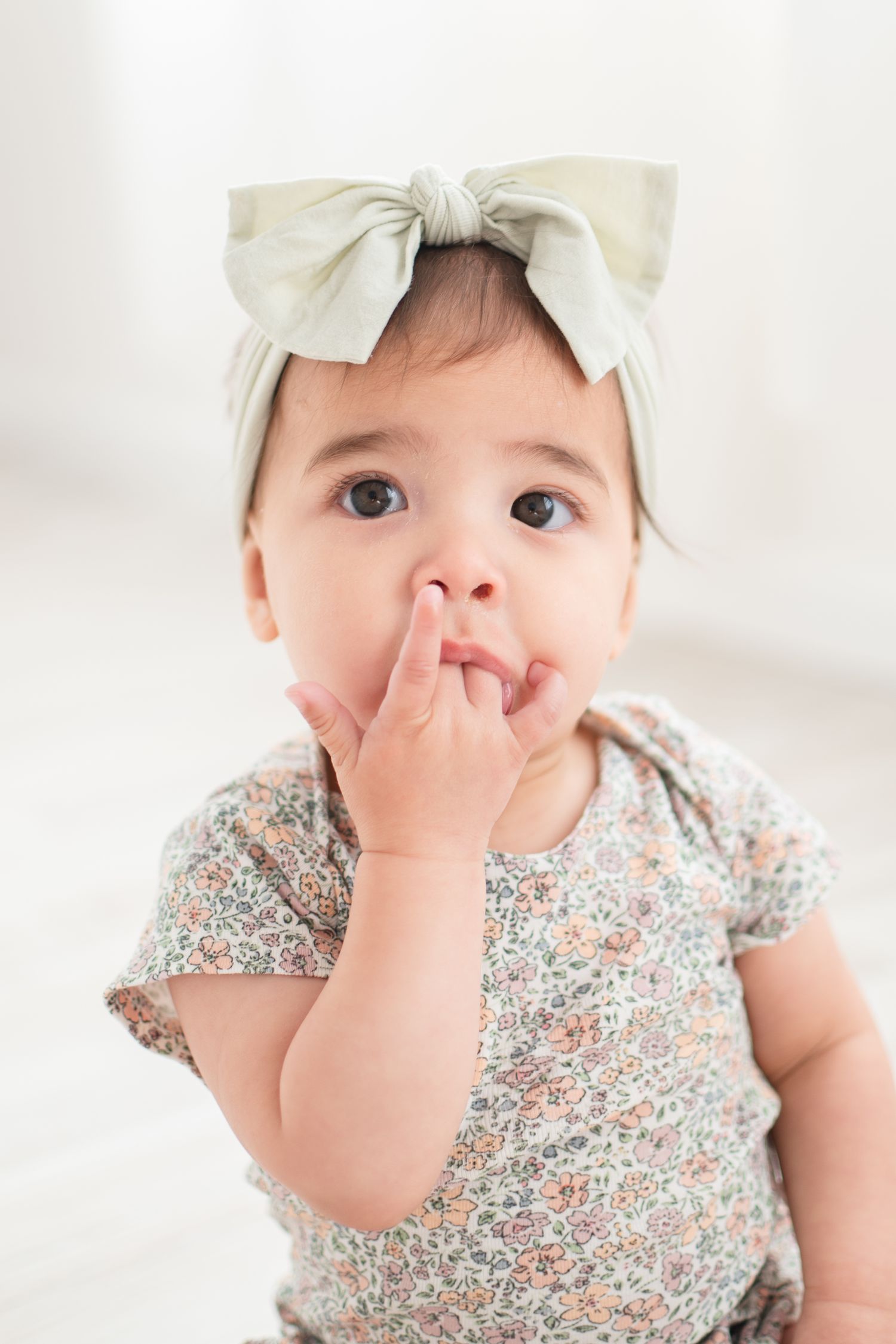 Sweet baby wearing floral print outfit and cream bow headband with finger near mouth in contemplative pose.
