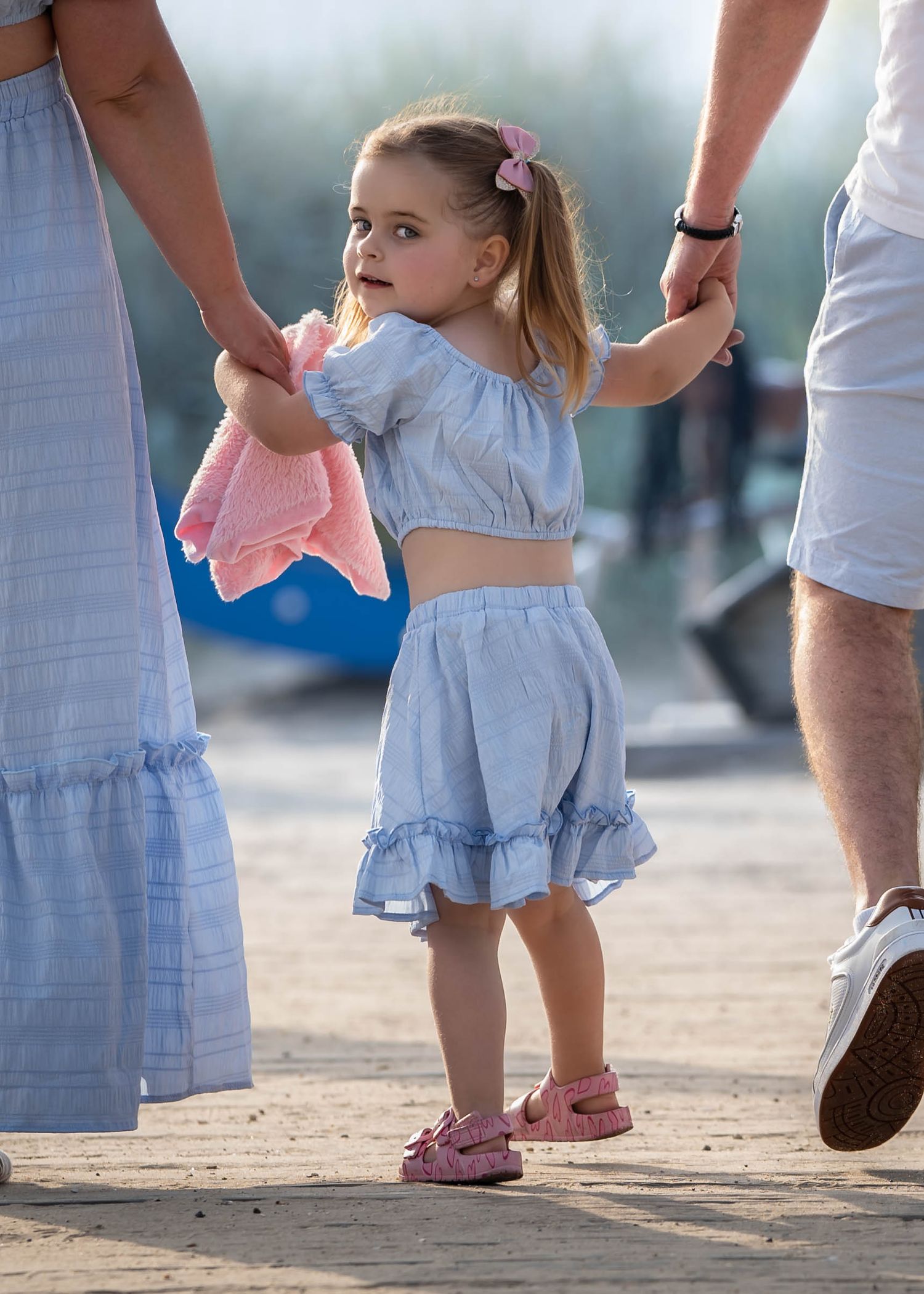 A small child in a ruffled blue two-piece outfit holds hands with adults while walking outdoors.