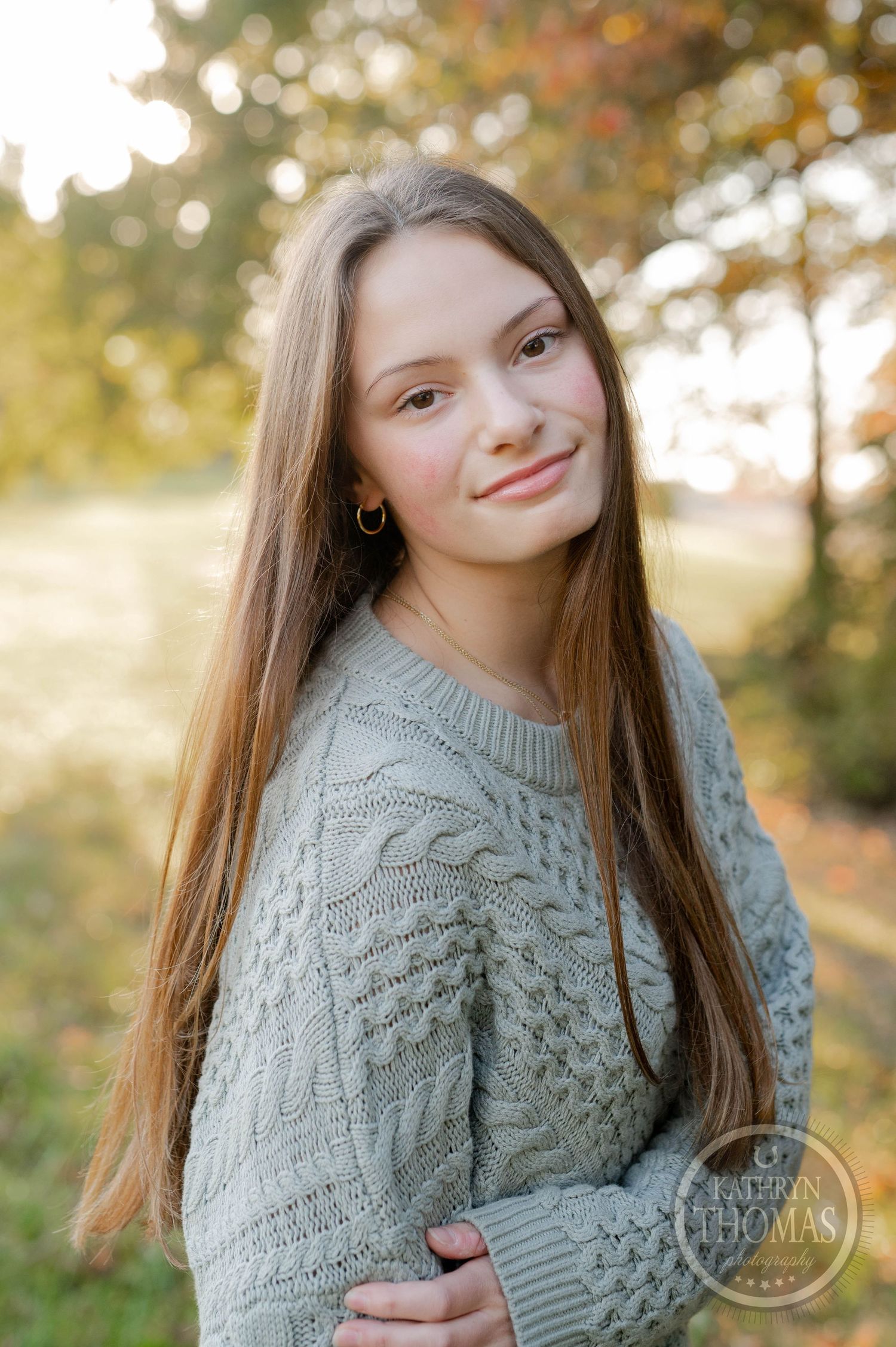A person in a gray cable knit sweater smiles while posing outdoors during autumn.