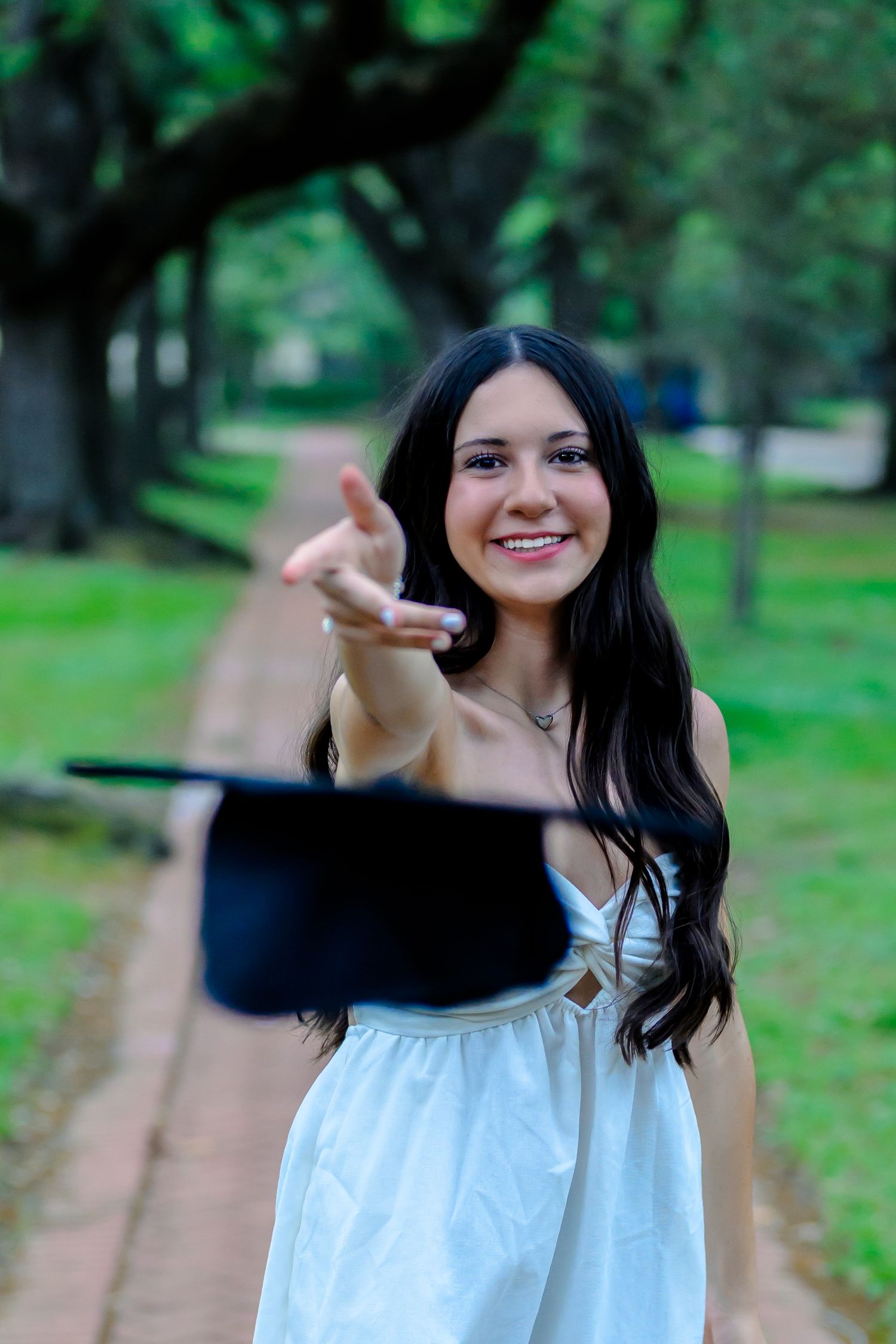 A smiling graduate in a white dress gestures while standing on a tree-lined path on graduation day.