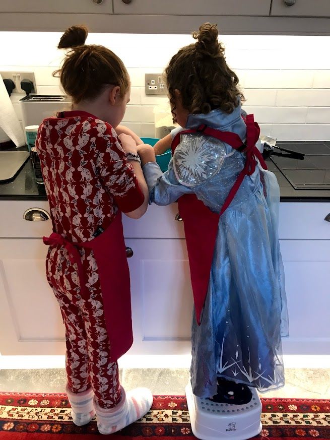 Two children in dresses stand together at a kitchen counter looking up at overhead cabinets.