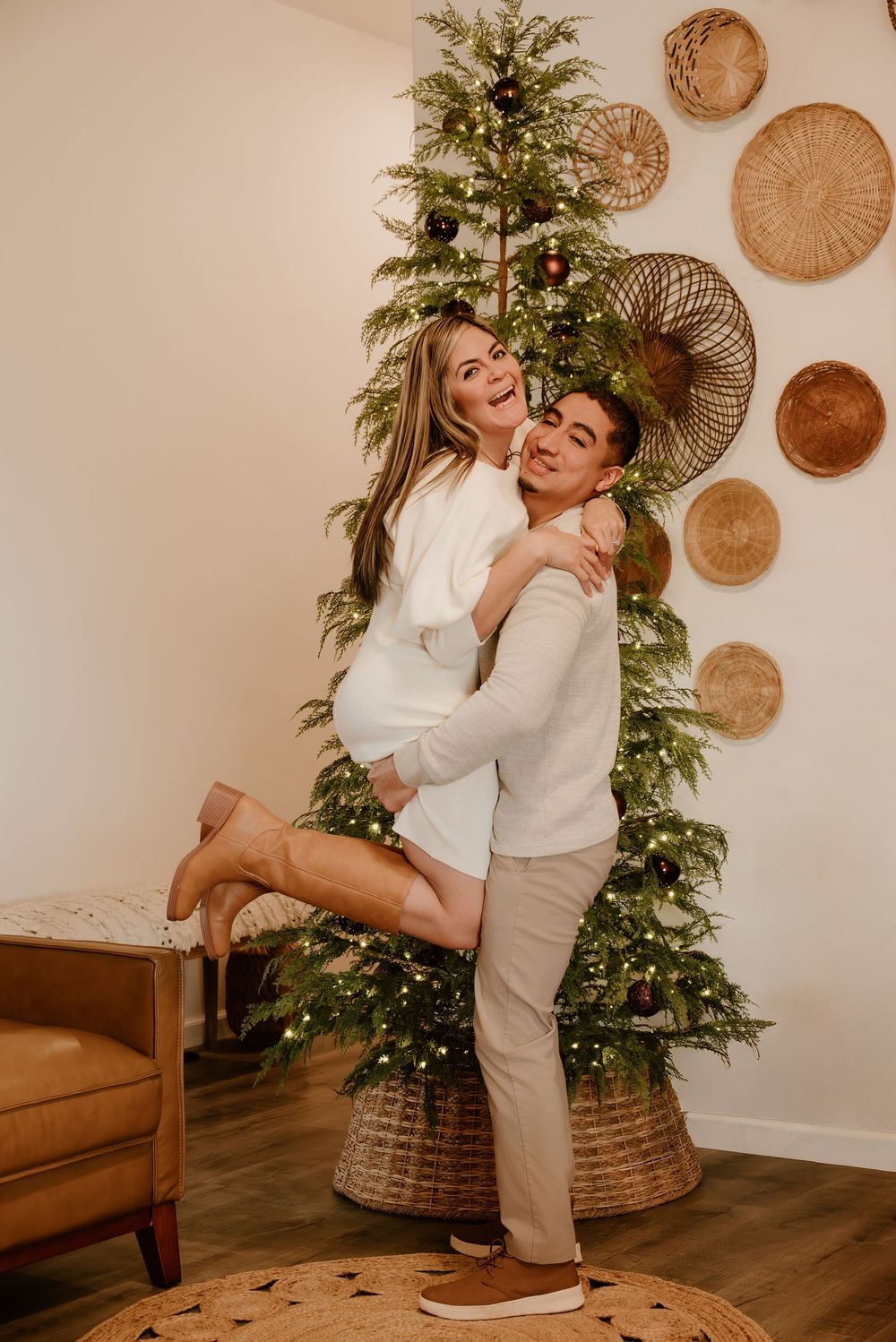 A couple embraces in front of a decorated Christmas tree with woven baskets on the wall in a cozy living room setting.