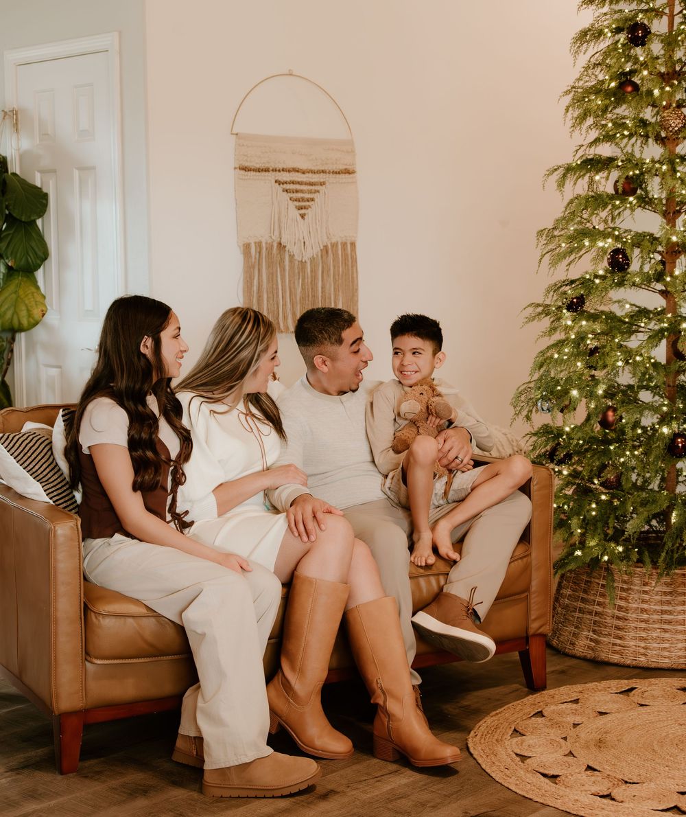 Family relaxes together on a brown leather couch next to a decorated Christmas tree in a cozy living room.