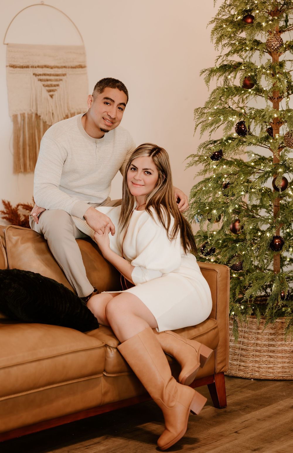 A couple sits together on a brown leather couch next to a decorated Christmas tree in a cozy living room setting.