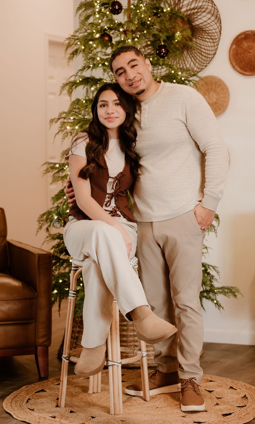 A couple poses in coordinated neutral outfits by a decorated Christmas tree in a cozy home setting.