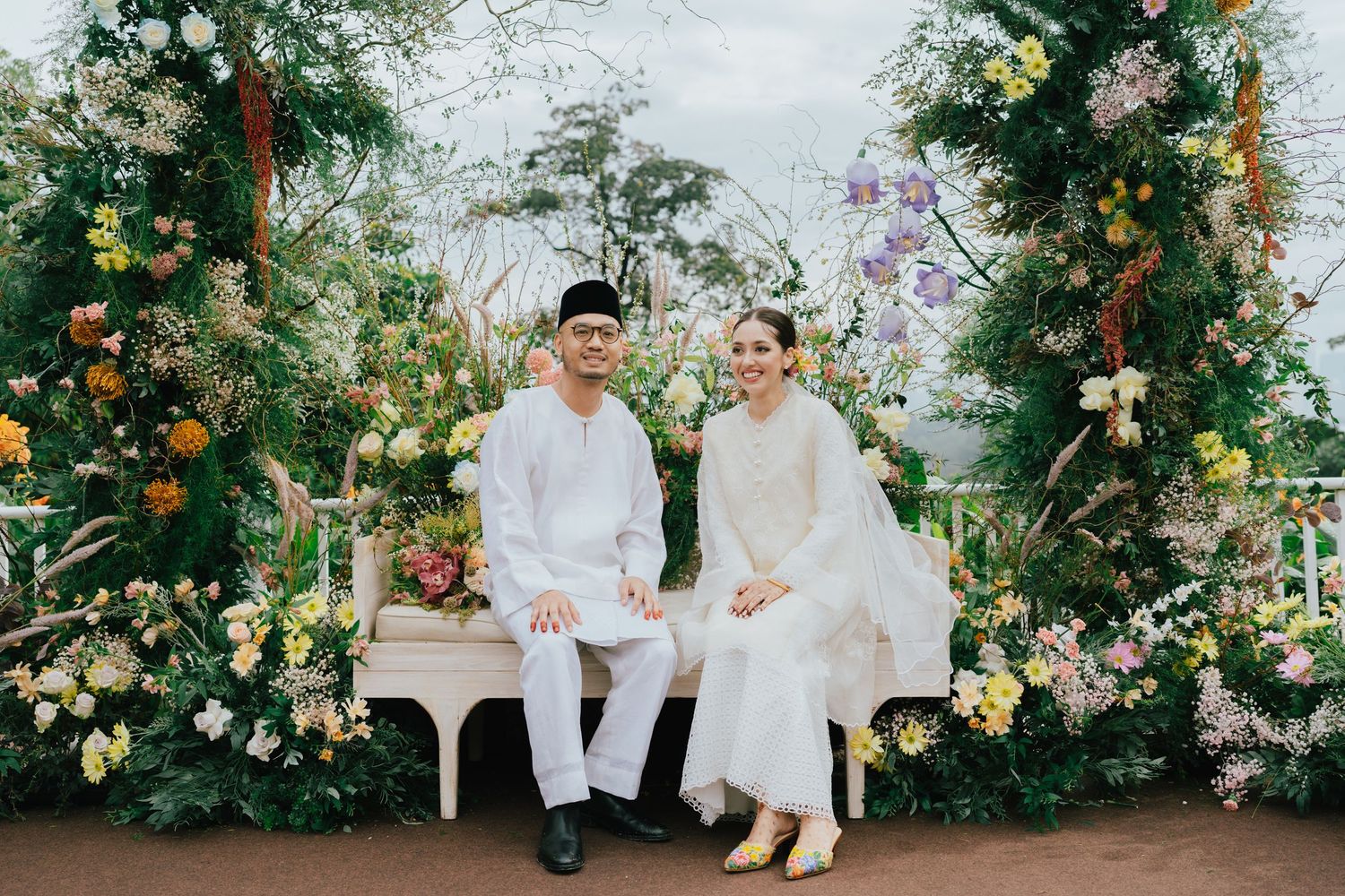 A couple in white traditional attire sits on a bench surrounded by lush floral arrangements at an elegant outdoor wedding.