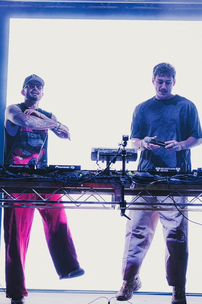DJs and electronic musicians perform with equipment on elevated stands in a backlit studio setting.