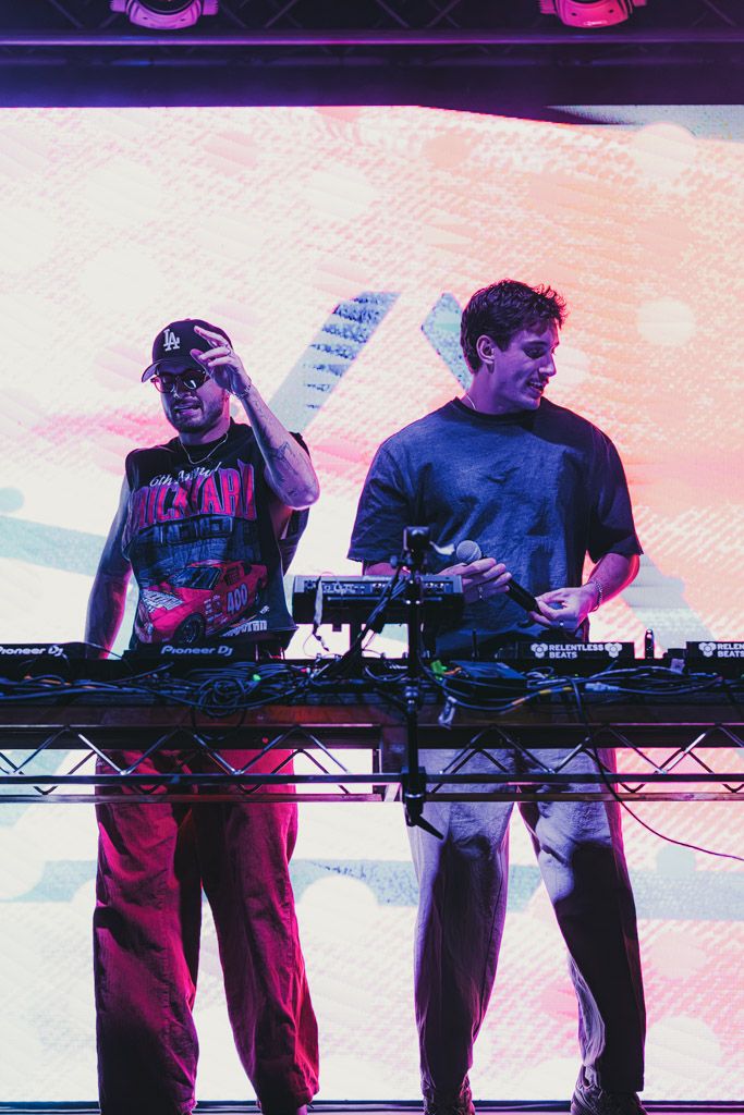 Electronic music performers play synthesizers and controllers on stage with pink and purple lighting effects.