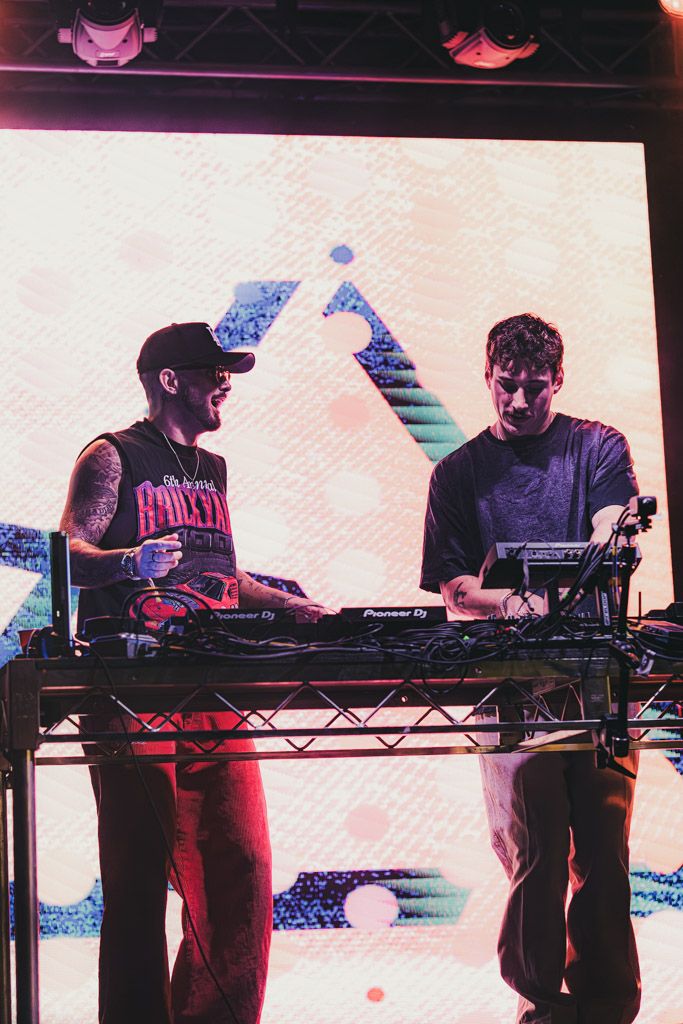 Two DJs perform on stage with electronic equipment under purple lighting at a music event.