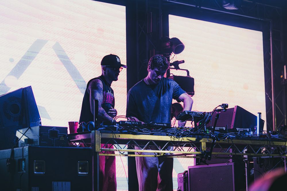 DJs performing at a concert with electronic equipment on stage under purple lighting effects.