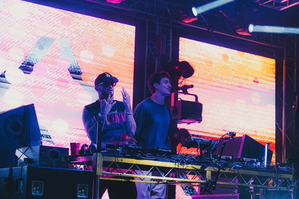 Colorful concert stage setup with vibrant pink and blue lighting illuminating DJ equipment and electronic displays.