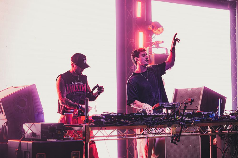 Electronic music performers on stage with bright purple and white backlighting creating a dramatic atmosphere.