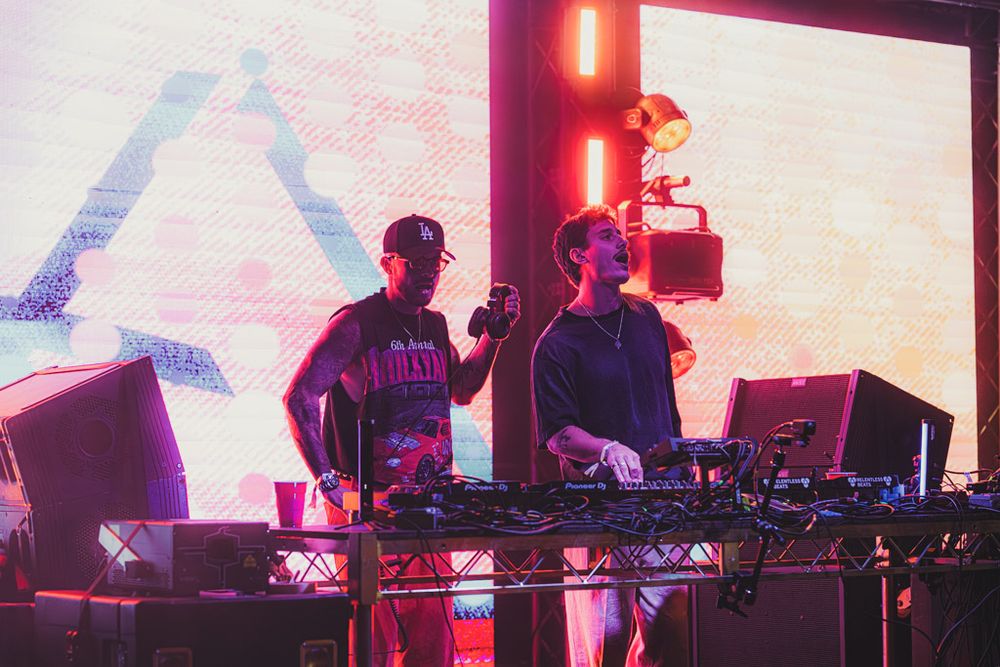 Electronic music performers on stage with synthesizers and equipment bathed in red and purple lighting effects.