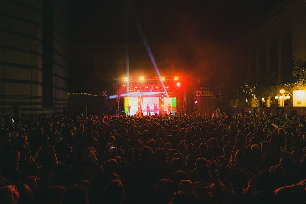 Large crowd of concert attendees watching illuminated stage with red lights at outdoor nighttime music performance.