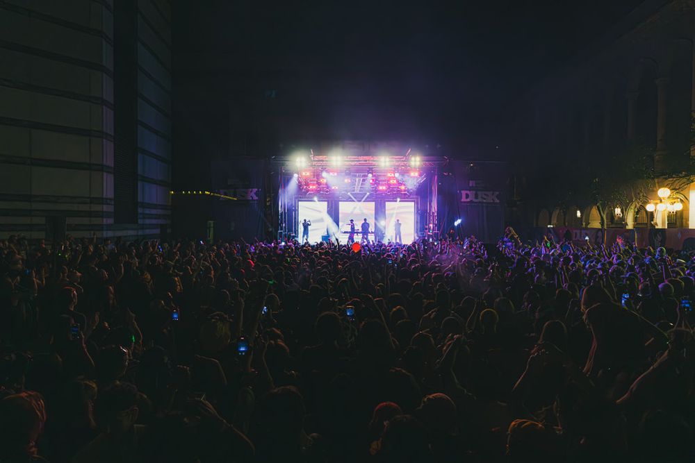 A large crowd of concert attendees stands before an illuminated stage with purple lighting at an outdoor nighttime event.