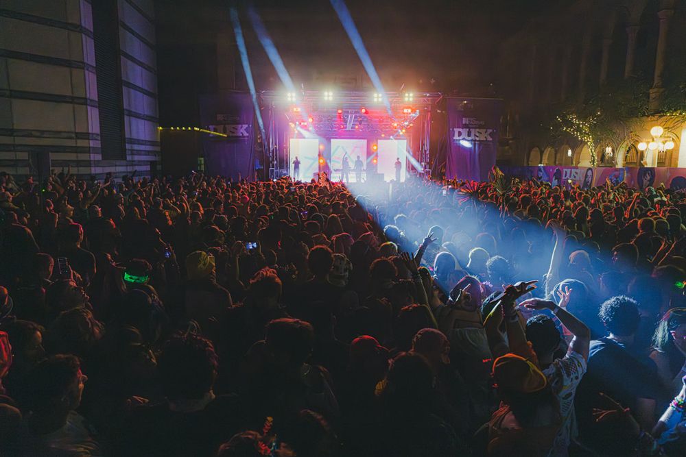 Large crowd at an electronic music concert with colorful purple and blue stage lighting illuminating the audience.